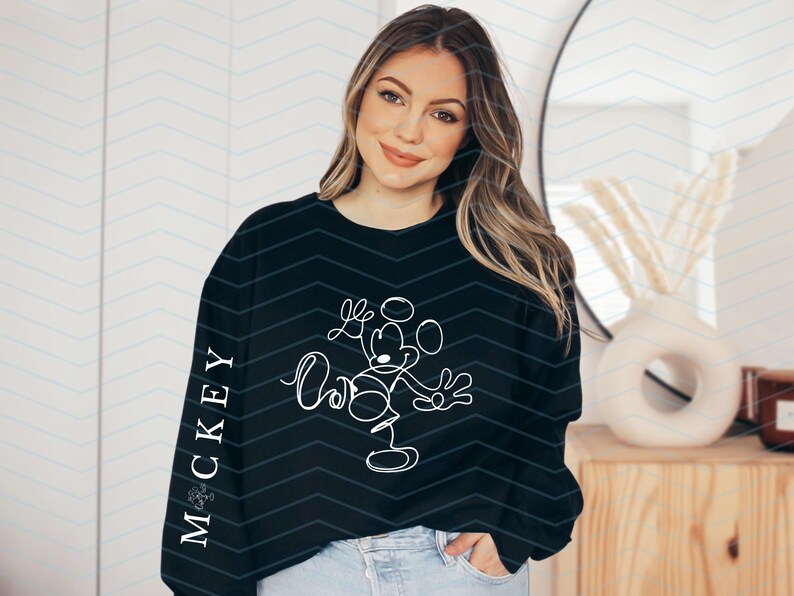 Puede incluir: Una sudadera negra de manga larga con un dibujo de l&iacute;nea continua blanca de Mickey Mouse en la parte delantera y el texto "MICKEY" impreso verticalmente en letras may&uacute;sculas blancas a lo largo del lado exterior de la manga derecha.