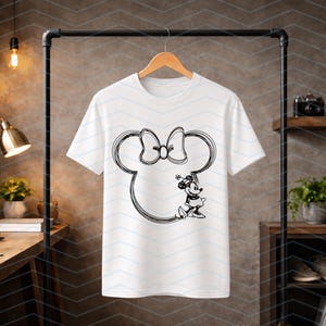 Puede incluir: Camiseta blanca con un contorno negro de la cabeza y el lazo de Minnie Mouse. El dise&ntilde;o incluye una ilustraci&oacute;n detallada de Minnie Mouse. La camiseta est&aacute; colgada en una percha de madera.