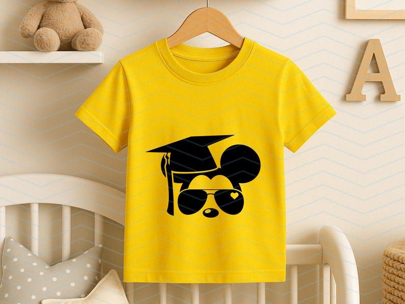 May include: Yellow t-shirt featuring a black graphic of a mouse wearing sunglasses and a graduation cap. The sunglasses have a heart shape on one lens. The shirt is hanging on a wooden hanger.