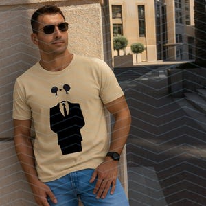May include: A light beige short-sleeved t-shirt featuring a black graphic print of a suit jacket, tie, and a stylized mouse-ear silhouette head. The shirt has a crew neckline and is made of a solid-colored fabric with a clean, simple design.