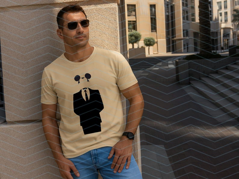 May include: A light beige short-sleeved t-shirt featuring a black graphic print of a suit jacket, tie, and a stylized mouse-ear silhouette head. The shirt has a crew neckline and is made of a solid-colored fabric with a clean, simple design.