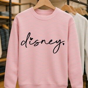 Op de afbeelding: Een lichtroze crewneck sweatshirt met het woord "disney" in zwart script. De "i" in het woord is vervangen door een Mickey Mouse-hoofd en een hart vervangt de punt. Andere sweatshirts hangen op de achtergrond.