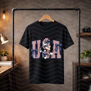 May include: A black short-sleeved t-shirt featuring a graphic of a cartoon mouse wearing a patriotic hat and outfit. The letters USA are printed in a red, white, and blue striped pattern behind the character, accented with blue bows.