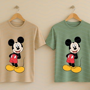 May include: Two short-sleeve crew neck t-shirts hanging on wooden hangers. One shirt is light tan and the other is sage green. Both feature a centered graphic print of Mickey Mouse wearing red shorts and yellow shoes on a white and black body.