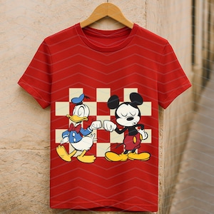 May include: Red t-shirt featuring Donald Duck and Mickey Mouse bumping fists, set against a checkered background. The shirt has a chevron pattern and is hanging on a wooden hanger. A classic cartoon design.