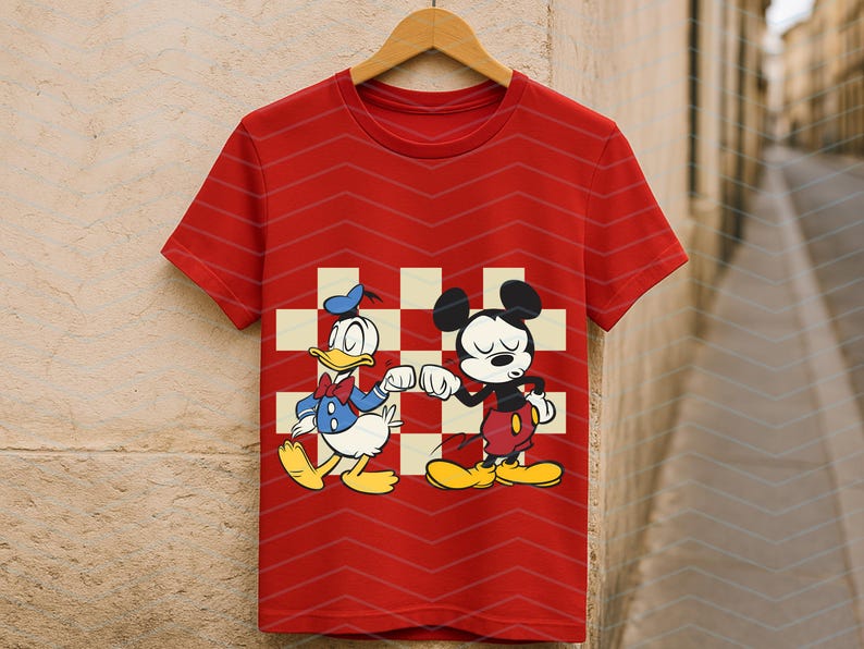 May include: Red t-shirt featuring Donald Duck and Mickey Mouse bumping fists, set against a checkered background. The shirt has a chevron pattern and is hanging on a wooden hanger. A classic cartoon design.