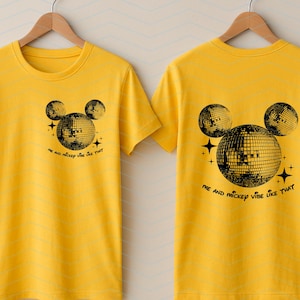 Puede incluir: Dos camisetas amarillas con una silueta negra de Mickey Mouse con orejas de bola de discoteca. El texto en las camisetas dice "Me and Mickey vibe like that."