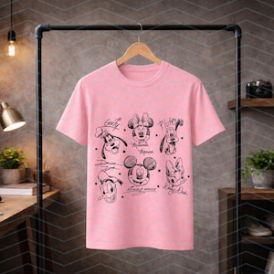 May include: A light pink t-shirt featuring black line drawings of Disney characters, including Mickey Mouse, Minnie Mouse, Goofy, Pluto, Donald Duck, and Daisy Duck. The names of the characters are written below their images.