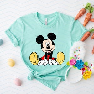 May include: A light blue t-shirt featuring a cartoon image of Mickey Mouse sitting down. Mickey Mouse is wearing red shorts and yellow shoes.