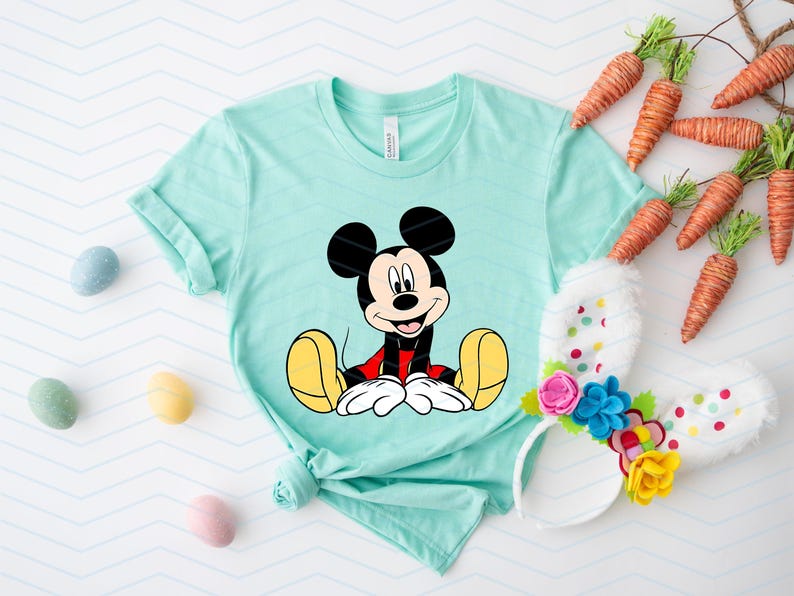 May include: A light blue t-shirt featuring a cartoon image of Mickey Mouse sitting down. Mickey Mouse is wearing red shorts and yellow shoes.