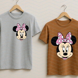 May include: Two t-shirts on wooden hangers. One is light gray, the other is brown. Each shirt features a Minnie Mouse graphic on the front. The graphic shows Minnie's face, black ears, and a pink bow with white polka dots.