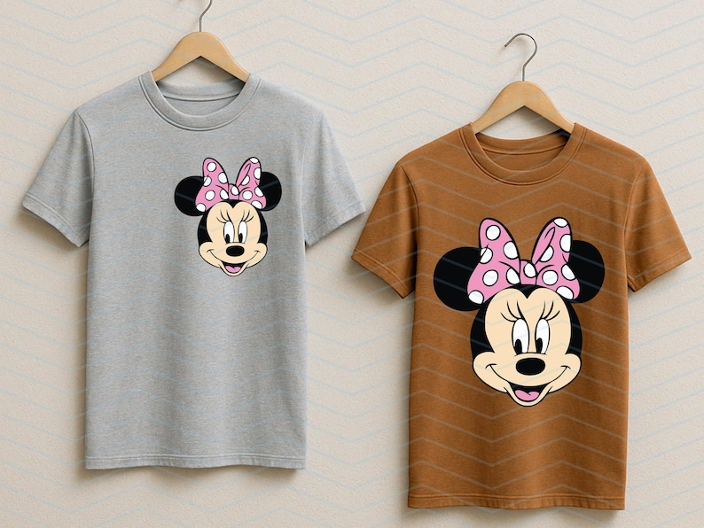 May include: Two t-shirts on wooden hangers. One is light gray, the other is brown. Each shirt features a Minnie Mouse graphic on the front. The graphic shows Minnie's face, black ears, and a pink bow with white polka dots.