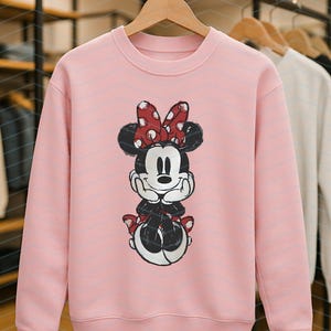 May include: A light pink sweatshirt featuring a Minnie Mouse graphic. The cartoon character is depicted with a red bow with white polka dots, black ears, and a black and white outfit. The sweatshirt is displayed on a wooden hanger.