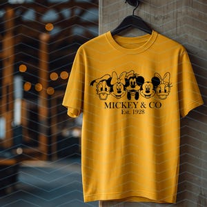 May include: A mustard yellow short-sleeve t-shirt featuring a black graphic print of five cartoon character faces in a row. Below the characters, the text reads MICKEY & CO and Est. 1928 in a black serif font. The shirt has a crew neckline.