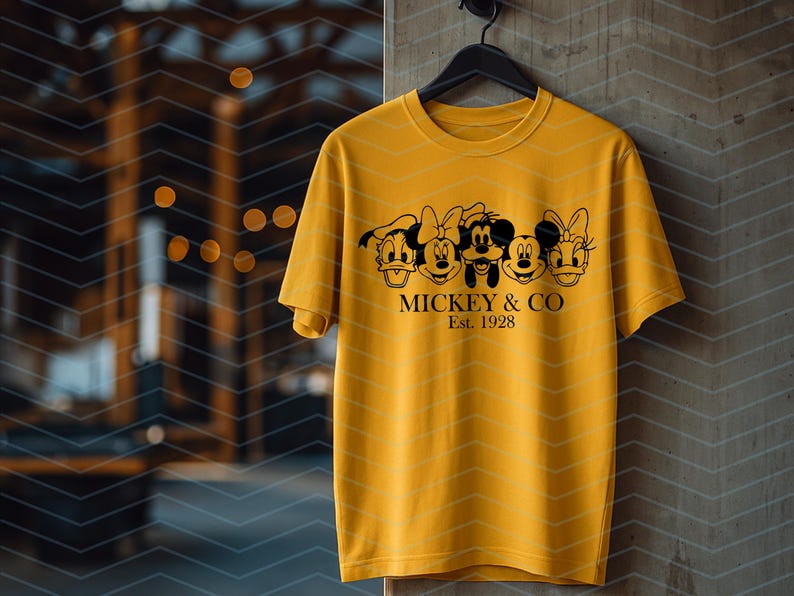 May include: A mustard yellow short-sleeve t-shirt featuring a black graphic print of five cartoon character faces in a row. Below the characters, the text reads MICKEY & CO and Est. 1928 in a black serif font. The shirt has a crew neckline.