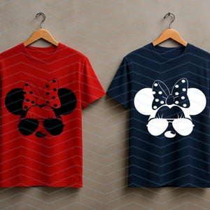 May include: Two t-shirts hanging on wooden hangers. One is red with a black and red polka-dot bow and sunglasses design. The other is navy blue with a white polka-dot bow and sunglasses design. Both shirts have a Minnie Mouse-inspired graphic.
