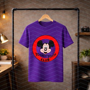 May include: A purple short-sleeved t-shirt featuring a graphic of Mickey Mouse inside a red circle. The text "Mickey Mouse Club" is printed in black lettering within the lower portion of the red circle. The shirt is displayed on a wooden hanger.