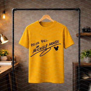 May include: A mustard yellow short-sleeve t-shirt featuring black text that reads Your Pal, Mickey Mouse, with a small black silhouette of Mickey Mouse ears integrated into the design, hanging on a wooden hanger against a neutral background.