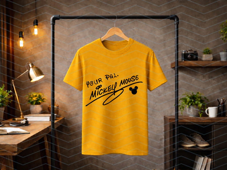 May include: A mustard yellow short-sleeve t-shirt featuring black text that reads Your Pal, Mickey Mouse, with a small black silhouette of Mickey Mouse ears integrated into the design, hanging on a wooden hanger against a neutral background.