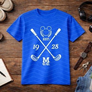 May include: Royal blue t-shirt with a white graphic featuring crossed golf clubs, a Mickey Mouse-inspired emblem, and the text "EST. 1928 M.C.C.". The shirt has a chevron pattern and is surrounded by accessories on a wooden surface.
