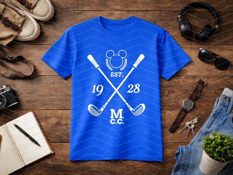 May include: Royal blue t-shirt with a white graphic featuring crossed golf clubs, a Mickey Mouse-inspired emblem, and the text "EST. 1928 M.C.C.". The shirt has a chevron pattern and is surrounded by accessories on a wooden surface.