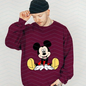 May include: A maroon sweatshirt featuring a cartoon image of Mickey Mouse sitting down. Mickey Mouse is wearing red shorts and yellow shoes.