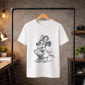 May include: White t-shirt featuring a black and white sketch of Minnie Mouse. The design includes Minnie's signature bow, polka-dot dress, and the text "Minnie Mouse xoxo". The shirt is hanging on a wooden hanger.