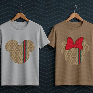 Puede incluir: Dos camisetas de manga corta y cuello redondo en perchas de madera. La camiseta de la izquierda es gris claro con una silueta de Mickey Mouse en tono beige con una franja roja y verde. La de la derecha es beige con un lazo rojo en la cabeza.