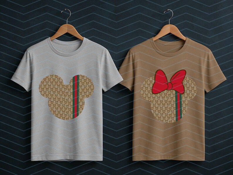 Puede incluir: Dos camisetas de manga corta y cuello redondo en perchas de madera. La camiseta de la izquierda es gris claro con una silueta de Mickey Mouse en tono beige con una franja roja y verde. La de la derecha es beige con un lazo rojo en la cabeza.