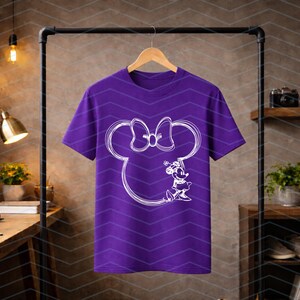 Puede incluir: Una camiseta morada con un contorno blanco de la cabeza y el lazo de Minnie Mouse, y una ilustraci&oacute;n detallada de Minnie Mouse. El dise&ntilde;o est&aacute; centrado en la camiseta.