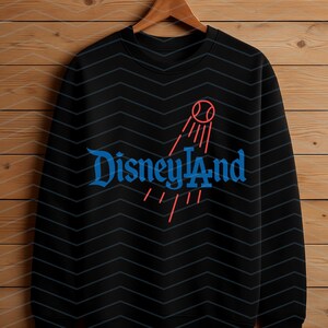 May include: Black crew neck sweatshirt featuring the word "Disneyland" in blue script, and a red graphic of a baseball with lines. The sweatshirt is displayed on a wooden hanger against a wood-panelled background.