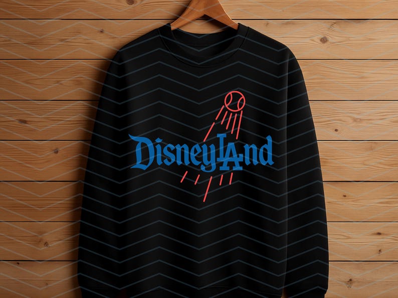 May include: Black crew neck sweatshirt featuring the word "Disneyland" in blue script, and a red graphic of a baseball with lines. The sweatshirt is displayed on a wooden hanger against a wood-panelled background.