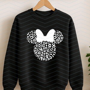 May include: Black sweatshirt featuring a white bow and a Minnie Mouse head design. The head and ears are filled with a black and white leopard print pattern. The sweatshirt is hanging on a wooden hanger.