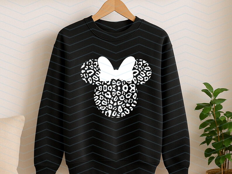 May include: Black sweatshirt featuring a white bow and a Minnie Mouse head design. The head and ears are filled with a black and white leopard print pattern. The sweatshirt is hanging on a wooden hanger.