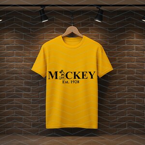 Pu&ograve; includere: Una maglietta gialla con la scritta "Mickey" in nero, con una grafica di Mickey Mouse e la scritta "Est. 1928". La maglietta &egrave; appesa a una gruccia di legno su uno sfondo di muro di mattoni.
