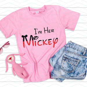 Op de afbeelding: Roze T-shirt met de tekst "I'm Her Mickey" in zwarte en rode letters. Het shirt ligt plat op een witte houten achtergrond met een zonnebril en een roze knuffelkonijn.