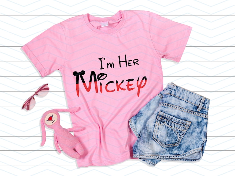 Op de afbeelding: Roze T-shirt met de tekst "I'm Her Mickey" in zwarte en rode letters. Het shirt ligt plat op een witte houten achtergrond met een zonnebril en een roze knuffelkonijn.