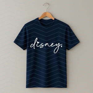Op de afbeelding: Marineblauw T-shirt met witte, cursieve "disney." tekst. Het shirt heeft een chevronpatroon en hangt aan een houten hanger. Een eenvoudig en stijlvol kledingstuk.