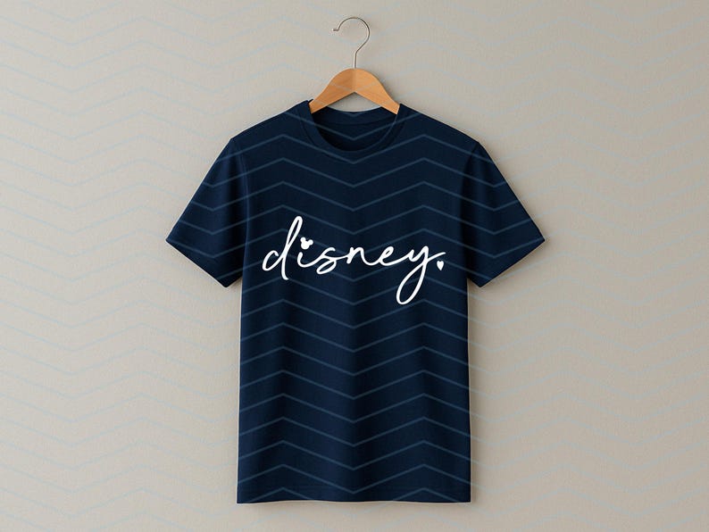 Op de afbeelding: Marineblauw T-shirt met witte, cursieve "disney." tekst. Het shirt heeft een chevronpatroon en hangt aan een houten hanger. Een eenvoudig en stijlvol kledingstuk.