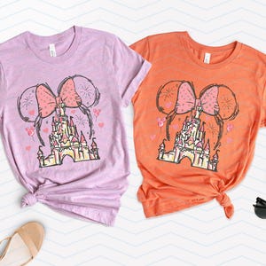 Pu&ograve; includere: Due magliette con uno schema di colori rosa e arancio che presentano un disegno animato del castello di Cenerentola con le orecchie di Minnie Mouse e un fiocco in cima.