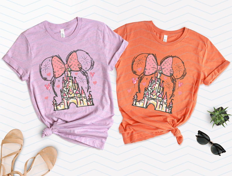 Pu&ograve; includere: Due magliette con uno schema di colori rosa e arancio che presentano un disegno animato del castello di Cenerentola con le orecchie di Minnie Mouse e un fiocco in cima.