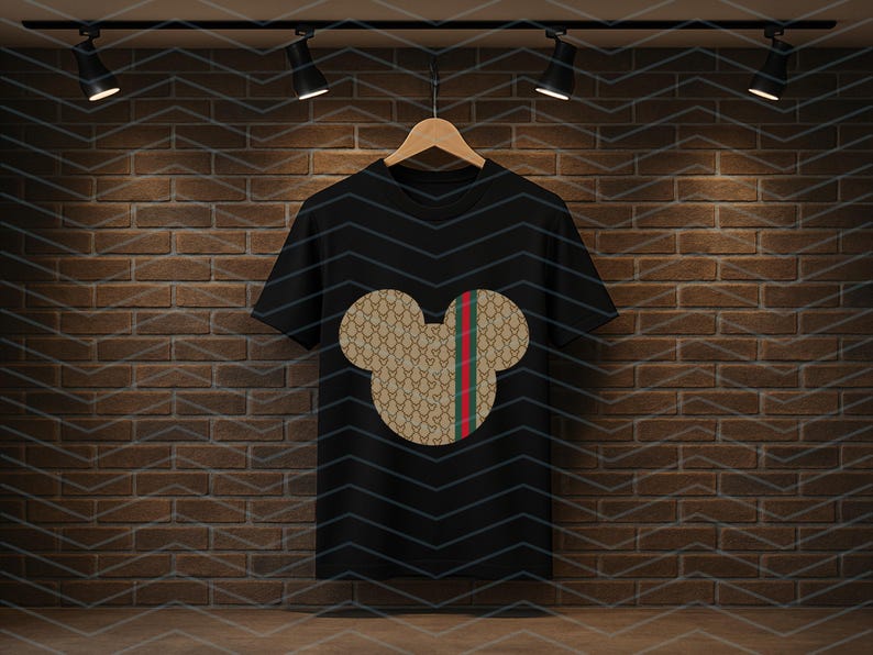 Puede incluir: Una camiseta negra de manga corta con un gr&aacute;fico central en forma de cabeza de rat&oacute;n. La silueta est&aacute; rellena con un patr&oacute;n repetitivo en tonos beige y marr&oacute;n e incluye una franja vertical de color rojo y verde en el lado derecho.