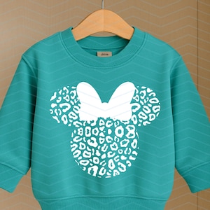 May include: Teal-colored sweatshirt featuring a white Minnie Mouse design with a leopard print pattern. The design includes a bow and two circular ears. The sweatshirt is hanging on a wooden hanger.