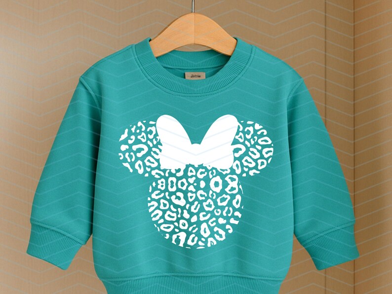 May include: Teal-colored sweatshirt featuring a white Minnie Mouse design with a leopard print pattern. The design includes a bow and two circular ears. The sweatshirt is hanging on a wooden hanger.