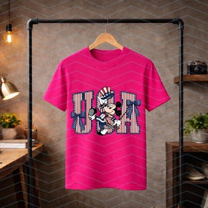 May include: A bright pink short-sleeved t-shirt featuring a large graphic on the front. The graphic displays the letters USA in a red and white striped pattern, accented with blue polka-dot bows, and a central illustration of Mickey Mouse.