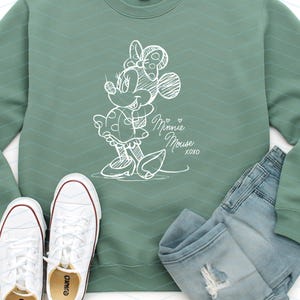 May include: Sage green sweatshirt with a white sketch of Minnie Mouse, paired with light wash denim jeans and white sneakers. The sweatshirt features the text "Minnie Mouse xoxo".