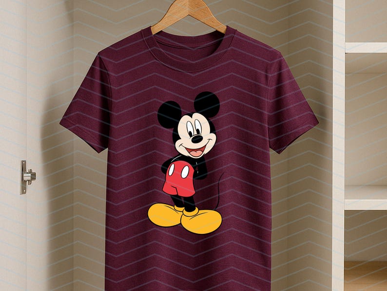 May include: A maroon short-sleeved t-shirt featuring a centered graphic of Mickey Mouse. The character is depicted with his signature black ears, red shorts, and large yellow shoes, standing with a cheerful expression on his face.