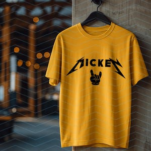 May include: Mustard yellow t-shirt with a black graphic that reads "MICKEY" in a stylized font, with a hand symbol. The shirt is hanging on a black hanger against a textured wall.
