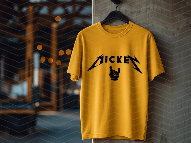 May include: Mustard yellow t-shirt with a black graphic that reads "MICKEY" in a stylized font, with a hand symbol. The shirt is hanging on a black hanger against a textured wall.