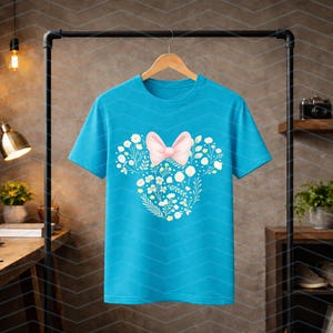 May include: A turquoise t-shirt with a floral design in the shape of a mouse head. The design includes a pink bow and white flowers. The shirt is hanging on a wooden hanger.